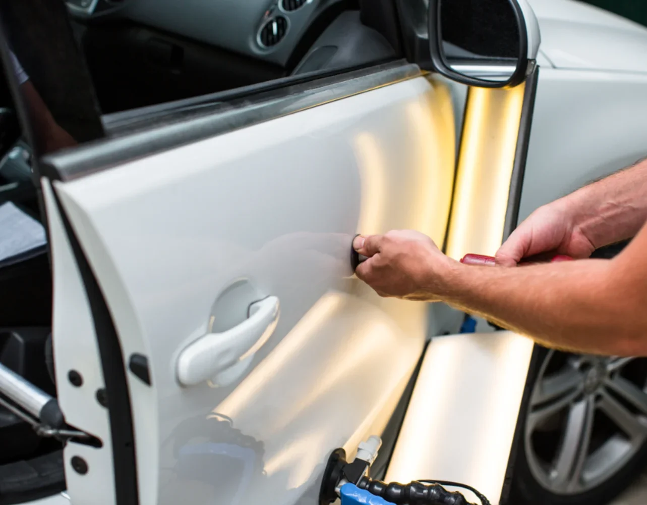 Technician using a dent-pulling tool on a white car door during panel beating repair