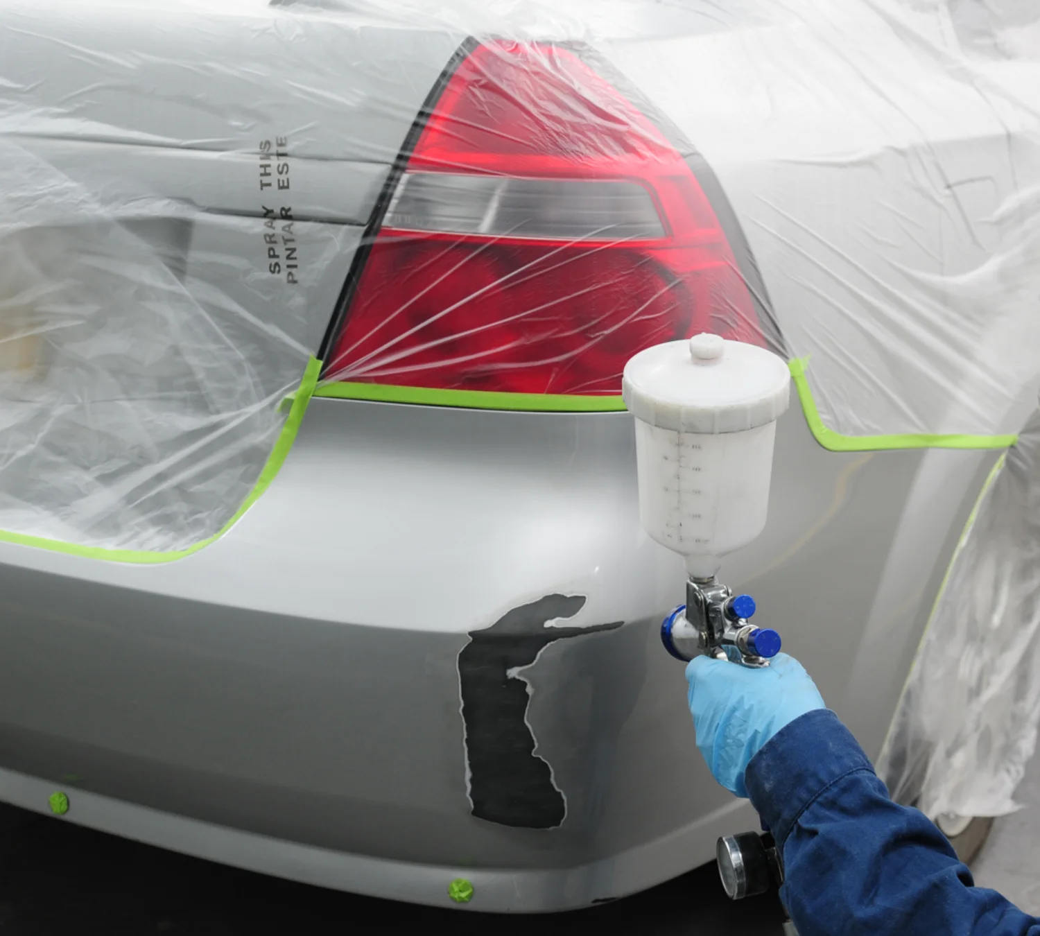 Spray painter applying paint to a masked car bumper during a panel refinishing job